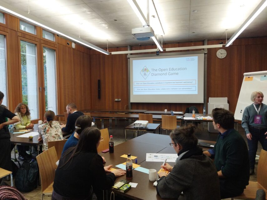 A seminar room with several groups of people sitting at tables and working together. In the background, a large projection screen shows a slide about the Open Education Diamond Game.